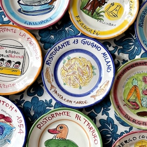Peut inclure: Ensemble d'assiettes en céramique peintes à la main, avec des motifs colorés et des noms de restaurants italiens. Chaque assiette présente des illustrations uniques, dont des poissons, des pâtes et un canard, avec du texte en italien. Elles sont disposées sur une nappe bleue à motifs floraux.