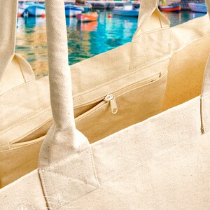 May include: A beige canvas tote bag with a zippered pocket and two long handles. The bag is made of natural cotton and has a simple, minimalist design.