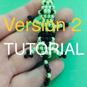 Puede incluir: Un llavero de lagarto de cuentas verdes y negras con el texto "Version 2 TUTORIAL" en una mano.