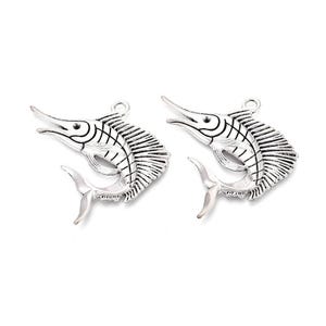May include: Two silver-colored marlin charms. Each charm features a detailed depiction of a marlin fish, with a long pointed bill, a dorsal fin, and a forked tail. The charms have a loop at the top for attaching to jewelry or other crafts.