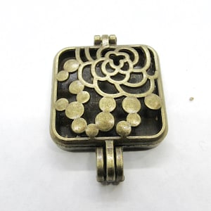 May include: A square-shaped, antique gold-toned metal locket with a delicate, openwork design featuring a stylised rose and a circle pattern. The locket has a hinged opening and a loop for attaching a chain.