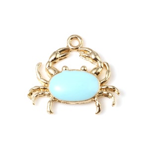 May include: A gold-coloured crab charm with a light blue oval centre. The crab has detailed claws and legs, and a small loop at the top for attaching to a necklace or bracelet. The charm is set against a white background.