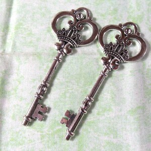 May include: Two silver-toned ornate skeleton keys with decorative crown-like tops. The keys have a vintage style and are perfect for crafting projects.