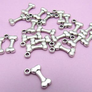 May include: A pile of silver-toned metal bone-shaped charms with a loop at the top. The charms are scattered on a light pink surface.
