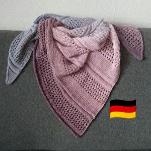 Może przedstawiać: Dzianinowy szal w odcieniach szarości, różu i fioletu. Trójkątny szal ma teksturowany wzór z ażurowymi detalami. Szal jest narzucony na szarą powierzchnię, prezentując gradient kolorów i skomplikowany wzór. Przytulny dodatek.