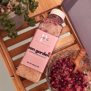 May include: A clear plastic bottle of Zen Garden handmade rose bath salts with a pink label, a glass bowl filled with bath salts and dried rose petals, a small wooden scoop, and a textured stone figure on a bamboo tray with a green succulent.