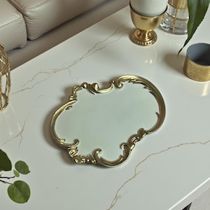 May include: A decorative tray with a pale green centre and gold ornate trim. The tray is on a white marble-effect surface. Other gold decorative items, including a birdcage and egg cup, are also visible, creating a luxurious aesthetic.