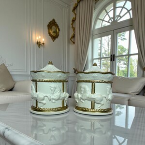 May include: Two white ceramic carousel-themed jars with gold detailing. Each jar has a carousel horse design and a gold-edged lid. The jars are displayed on a marble surface in a well-lit room.