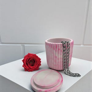 May include: A red rose, a pink and white ceramic container with a silver chain, and a matching lid are arranged on a white surface. The container has vertical ridges and a marbled effect.