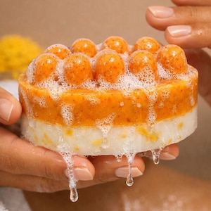 May include: A round, two-toned massage soap bar featuring a textured orange top layer with raised, rounded nodes and a smooth white bottom layer. The soap is covered in white lather and bubbles that are dripping down the sides of the bar.