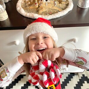 May include: A young child wearing a Santa hat and pajamas holds a red bucket filled with candy canes. The bucket has a white trim and a black belt with a gold buckle. The child is smiling and looking at the camera.