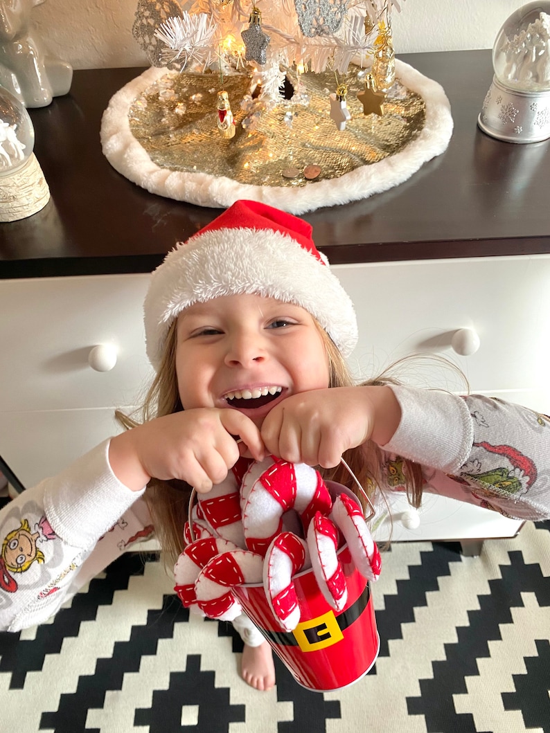May include: A young child wearing a Santa hat and pajamas holds a red bucket filled with candy canes. The bucket has a white trim and a black belt with a gold buckle. The child is smiling and looking at the camera.