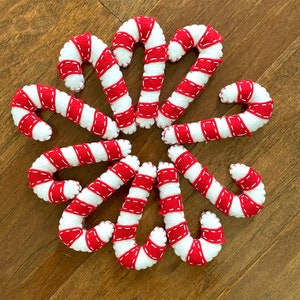 May include: A collection of ten felt candy canes in red and white with white stitching. The candy canes are arranged in a circle on a wood surface.