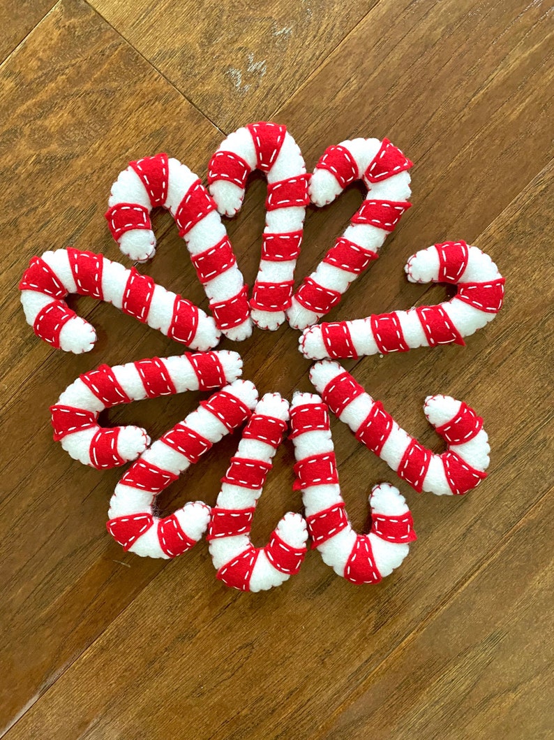 May include: A collection of ten felt candy canes in red and white with white stitching. The candy canes are arranged in a circle on a wood surface.