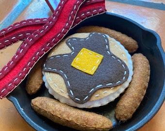 Könnte beinhalten: Ein Filzpfannkuchen mit braunem Sirup und einer gelben Butterflocke, umgeben von vier Filzwurststücken in einer schwarzen gusseisernen Pfanne mit rotem Griff. Zwei Filzstreifen Speck liegen auf der Pfanne.