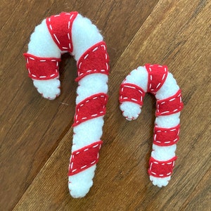 May include: Two felt candy canes, one larger than the other, with red and white stripes. The candy canes are on a wood background.