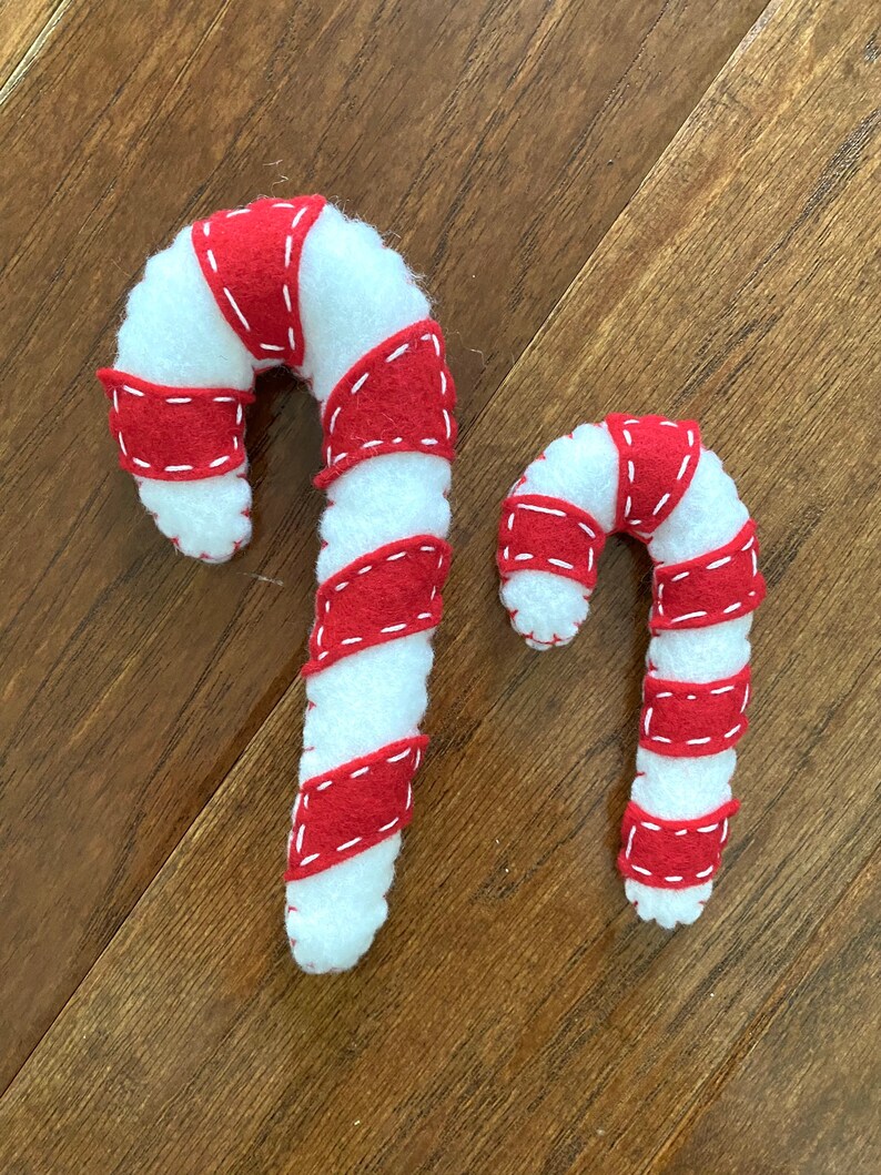 May include: Two felt candy canes, one larger than the other, with red and white stripes. The candy canes are on a wood background.