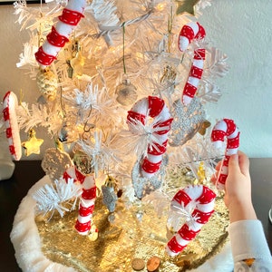 May include: A white artificial Christmas tree decorated with red and white candy cane ornaments, gold stars, and other festive decorations. The tree is sitting on a gold sequin tree skirt with a white fur trim.