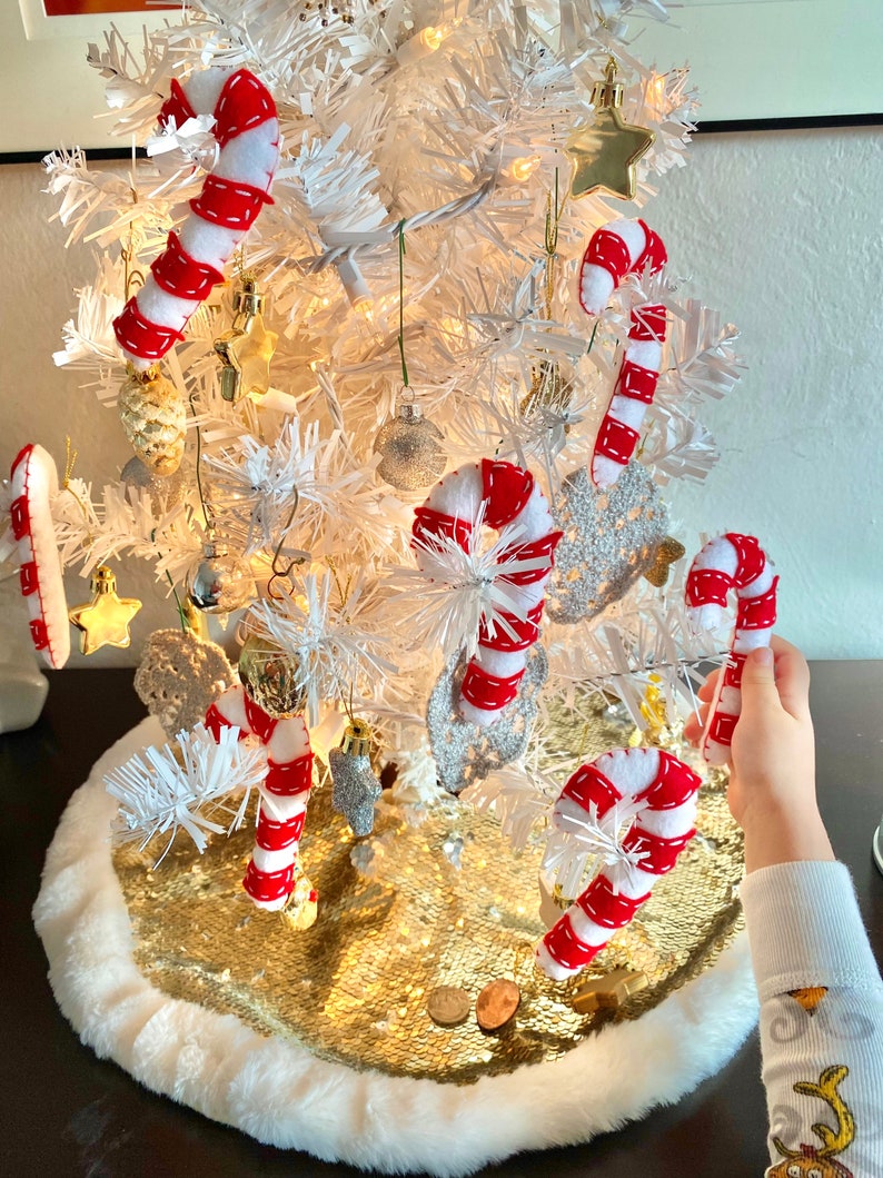 May include: A white artificial Christmas tree decorated with red and white candy cane ornaments, gold stars, and other festive decorations. The tree is sitting on a gold sequin tree skirt with a white fur trim.