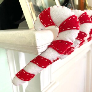 May include: A red and white candy cane ornament with white felt and red felt stripes, sewn together with white thread. The candy cane is hanging on a white mantel.