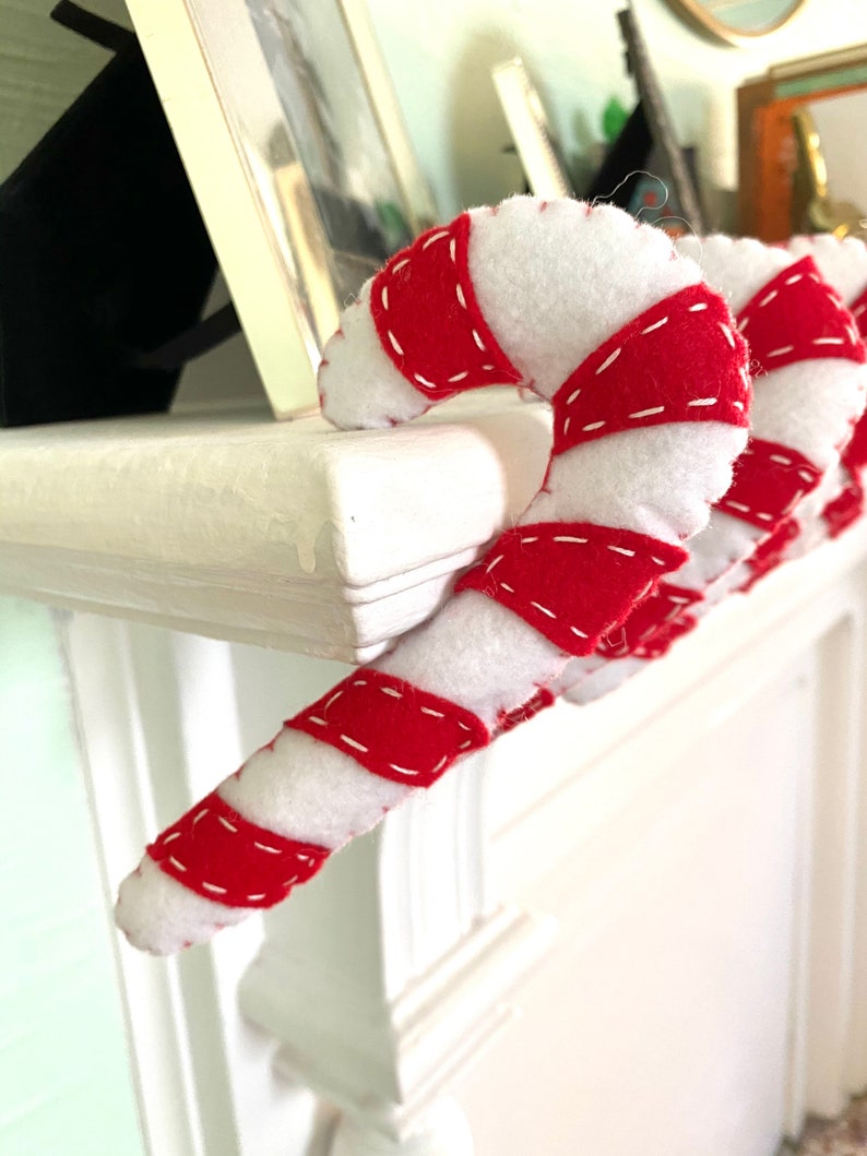May include: A red and white candy cane ornament with white felt and red felt stripes, sewn together with white thread. The candy cane is hanging on a white mantel.