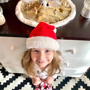 May include: A young girl wearing a red Santa hat and a white shirt with a Christmas-themed print is sitting on a black and white checkered floor. She is looking up at the camera with a smile.