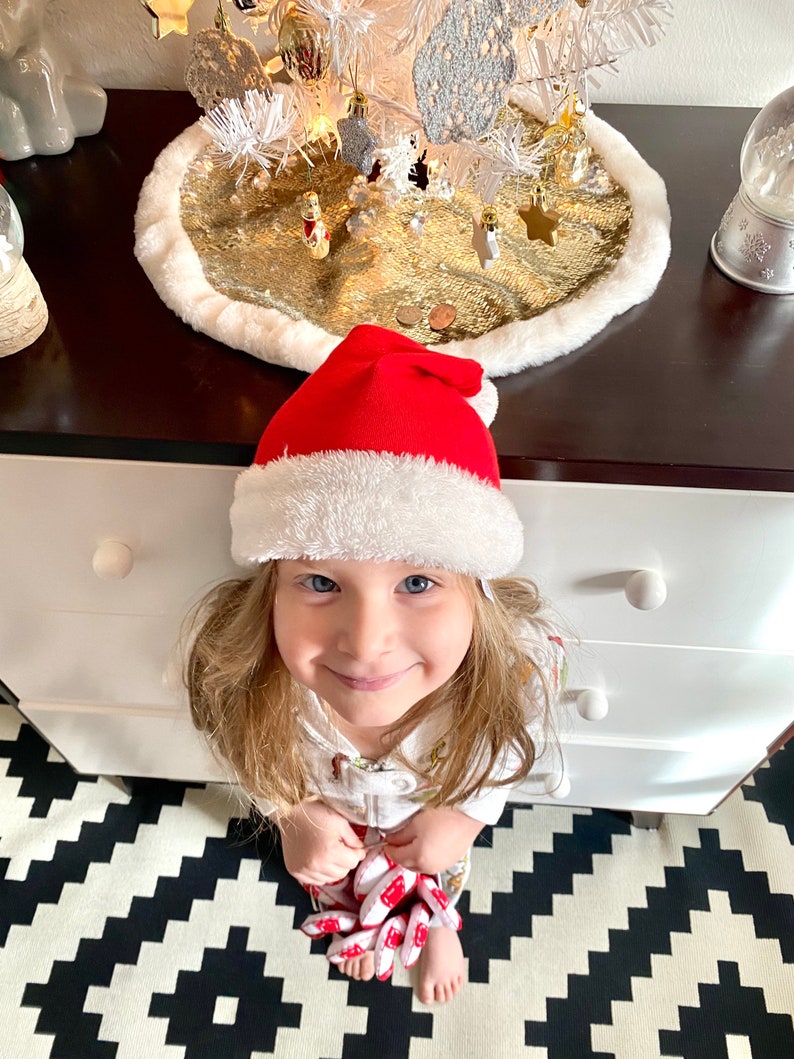 May include: A young girl wearing a red Santa hat and a white shirt with a Christmas-themed print is sitting on a black and white checkered floor. She is looking up at the camera with a smile.