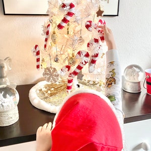 May include: A child wearing a red Santa hat decorates a white artificial Christmas tree with candy cane ornaments. The tree is decorated with gold and white ornaments and lights.