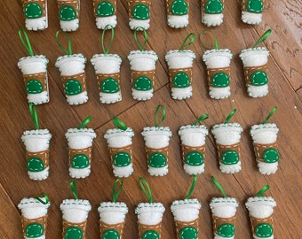 May include: Thirty-six felt coffee cup ornaments with green and brown details. The cups are white with a brown band and a green lid. Each cup has a green and white stitched design.