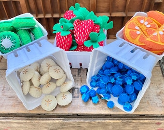 Puede incluir: Una colección de juguetes de frutas de fieltro, que incluye fresas, kiwis, naranjas, plátanos y arándanos, exhibidos en recipientes de papel blanco. Las fresas son rojas con tapas verdes, los kiwis son verdes con centros blancos y las naranjas son naranjas. Los plátanos son de color canela y los arándanos son azules.