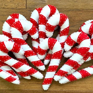 May include: A group of ten felt candy canes in red and white with white stitching. The candy canes are arranged in a cluster on a wood surface.
