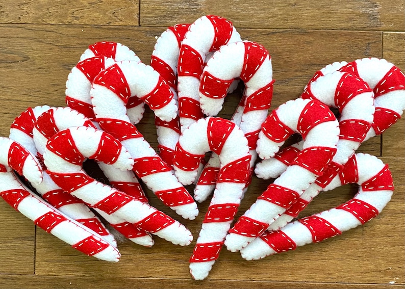 May include: A group of ten felt candy canes in red and white with white stitching. The candy canes are arranged in a cluster on a wood surface.