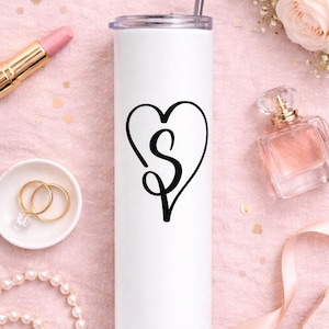 May include: White insulated tumbler with a black heart-shaped design containing the letter "S". The tumbler has a silver straw and lid. The background includes a pink surface, lipstick, rings, perfume, and flowers.