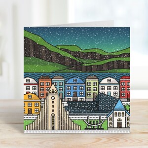 Op de afbeelding: Een gevouwen wenskaart met een kleurrijke illustratie van een stadsgezicht onder een sterrenhemel. Het ontwerp omvat gebouwen met verschillende gekleurde daken, een kerk en een bergketen. De kaart ligt op een houten oppervlak.