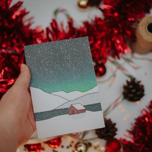 May include: A hand holds a Christmas card featuring a snowy landscape with a red cabin under a starry sky. The card is surrounded by red tinsel, ornaments, and a pine cone, suggesting a festive theme.