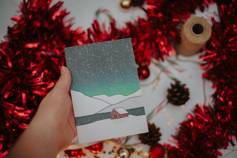 May include: A hand holds a Christmas card featuring a snowy landscape with a red cabin under a starry sky. The card is surrounded by red tinsel, ornaments, and a pine cone, suggesting a festive theme.
