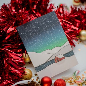 May include: A Christmas card featuring a winter landscape with a starry night sky, snow-covered hills, and a small red cabin. The card is surrounded by red tinsel, gold and red ornaments, and festive decorations.