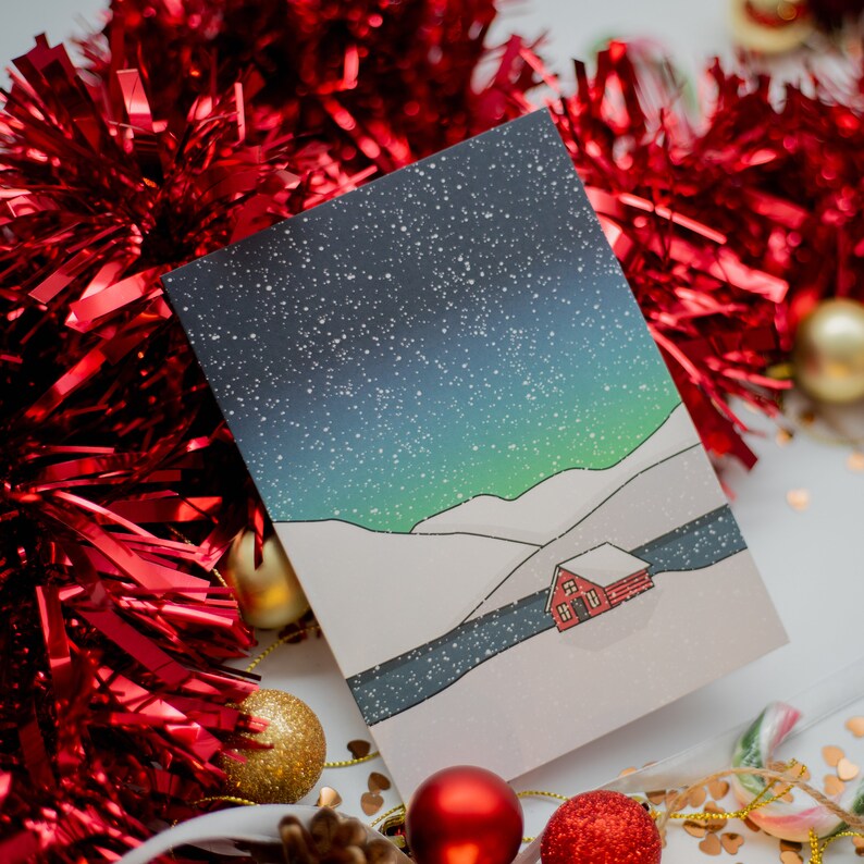 May include: A Christmas card featuring a winter landscape with a starry night sky, snow-covered hills, and a small red cabin. The card is surrounded by red tinsel, gold and red ornaments, and festive decorations.