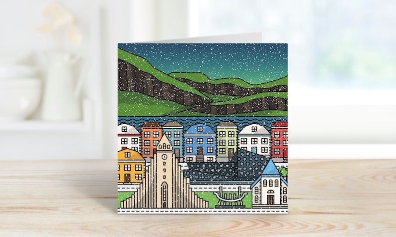 Op de afbeelding: Een gevouwen wenskaart met een kleurrijke illustratie van een stadsgezicht onder een sterrenhemel. Het ontwerp omvat gebouwen met verschillende gekleurde daken, een kerk en een bergketen. De kaart ligt op een houten oppervlak.
