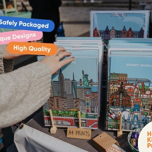 May include: A display of colorful city skyline prints, including Disney World, Amsterdam, and New York City. The prints are on stands and labeled "A4 Prints". The text "Hannah Kirkland Prints" is also visible.