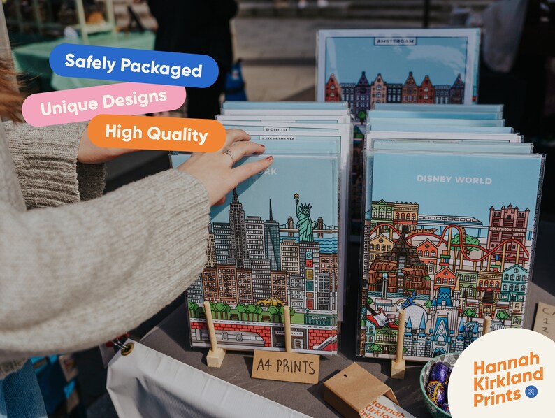 May include: A display of colorful city skyline prints, including Disney World, Amsterdam, and New York City. The prints are on stands and labeled "A4 Prints". The text "Hannah Kirkland Prints" is also visible.