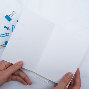 May include: A blank, white greeting card is held open, revealing its interior. Scattered around are blue binder clips, paper clips, and push pins. The items rest on a white and gray marble surface, creating a clean, organized aesthetic.