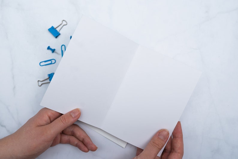 May include: A blank, white greeting card is held open, revealing its interior. Scattered around are blue binder clips, paper clips, and push pins. The items rest on a white and gray marble surface, creating a clean, organized aesthetic.