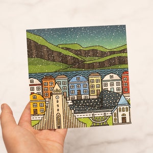 Op de afbeelding: Een kleurrijke illustratie van een stadsgezicht met huizen, een kerk en bergen onder een sterrenhemel. De huizen zijn in verschillende kleuren, waaronder geel, blauw en oranje. Het kunstwerk heeft een getextureerde, vintage uitstraling.