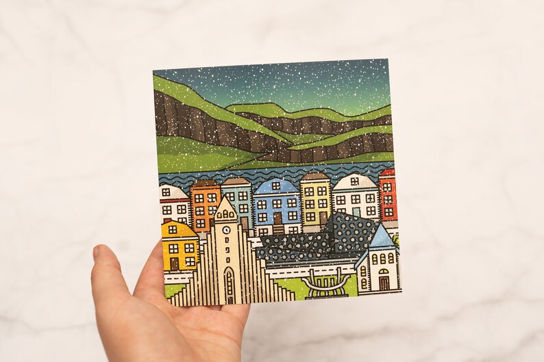 Op de afbeelding: Een kleurrijke illustratie van een stadsgezicht met huizen, een kerk en bergen onder een sterrenhemel. De huizen zijn in verschillende kleuren, waaronder geel, blauw en oranje. Het kunstwerk heeft een getextureerde, vintage uitstraling.
