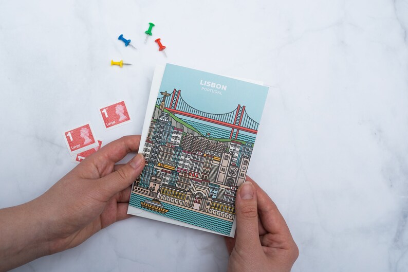 May include: A postcard featuring a detailed illustration of Lisbon, Portugal, with a bridge and buildings. The postcard is held in hands, with red stamps and colorful push pins nearby. The text "LISBON PORTUGAL" is at the top.