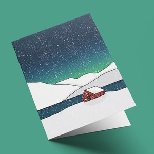 May include: A folded greeting card featuring a winter landscape illustration. The design shows a red cabin nestled in a snowy valley under a starry night sky with green aurora borealis. The card is on a teal background.