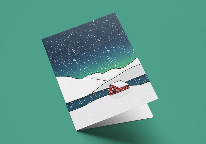 May include: A folded greeting card featuring a winter landscape illustration. The design shows a red cabin nestled in a snowy valley under a starry night sky with green aurora borealis. The card is on a teal background.