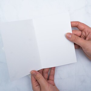 May include: A blank, white, folded card is held open by two hands against a marble-patterned background. The card is centered in the frame, with the hands positioned on either side, displaying the inside of the card.
