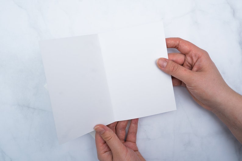 May include: A blank, white, folded card is held open by two hands against a marble-patterned background. The card is centered in the frame, with the hands positioned on either side, displaying the inside of the card.